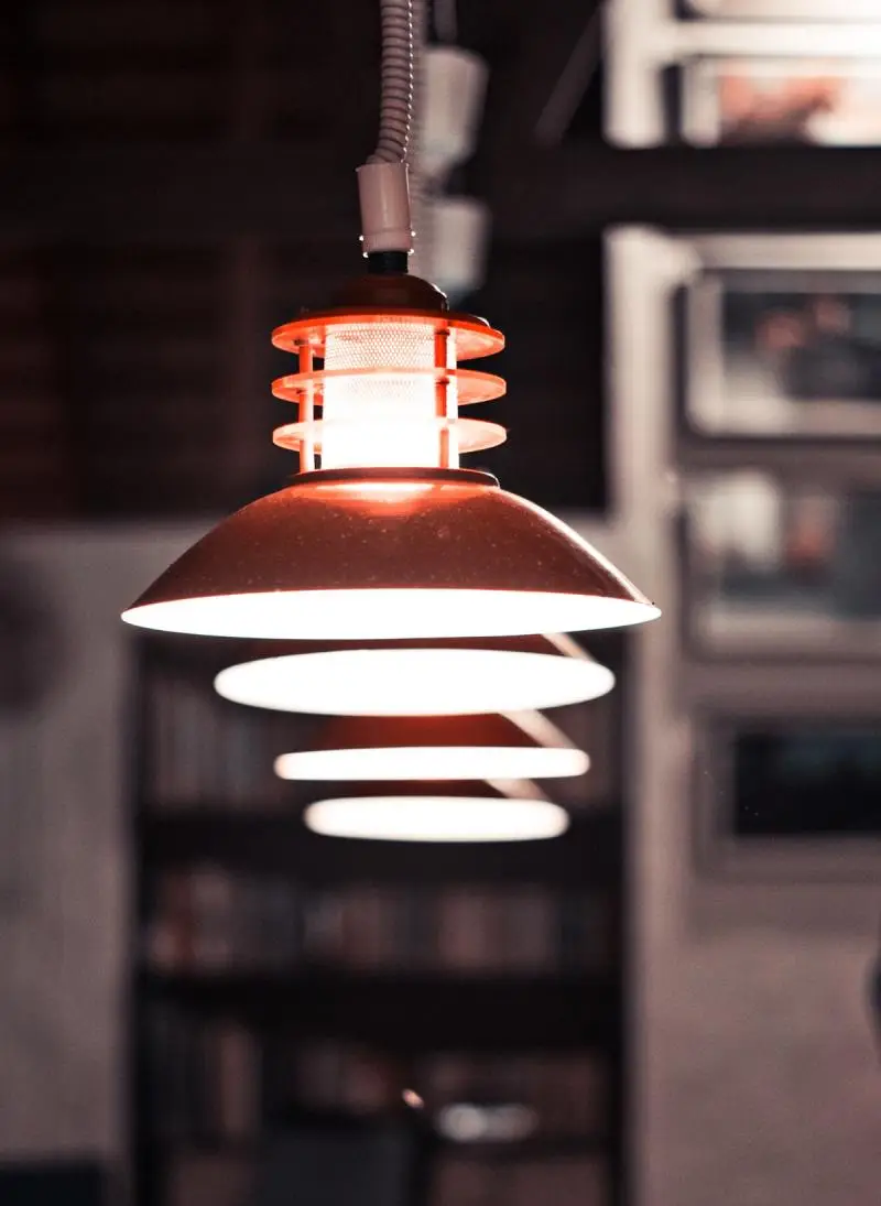 A stylish, hanging lamp with a red shade and layered design, emitting a warm light, set against a blurred background of shelves.